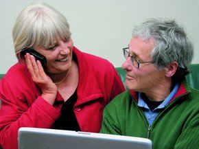 Starší pár před notebookem se vzájemně na sebe dívá, zatímco žena mluví do telefonu